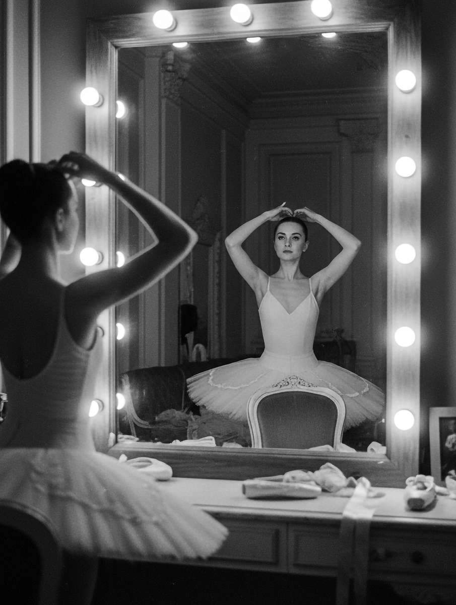 Dancer on pointe captured mid-balance facing the mirror, fingers extended and eyes fixed on her own reflection for precise…
